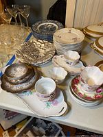 Overview of various plates, teacups, and saucers including floral and historic colonial scene patterns arranged on a table.