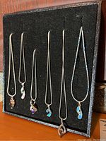 Front view of six necklaces with opal-like and colored stone pendants arranged on black felt display, showing variety of pendant designs and chain types.