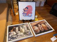 Overall view of three framed prints on the floor, showing the print by Barbara Lavallee and two food prints in white frames, one with separated joint.
