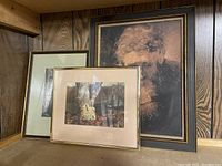 Three framed art prints arranged leaning against wood paneled wall: large portrait, middle sized nature print, smaller framed art.
