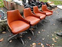 Four mid-century modern swivel chairs with bent wood sides and orange vinyl upholstery lined up outside on pavement with leaves and background litter and vehicles.