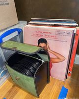 Side view of vintage 45 RPM record carrying case with lid open showing stacked vinyl records inside. Background stack of visible record sleeves including Lena Horne at Waldorf Astoria.
