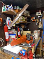 Photo showing shelves and pegboard with Husqvarna chainsaw with orange cover, fire extinguisher, caulking tubes, boxes, paint brushes and other workshop hardware items.
