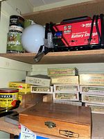 Shelf with Husky battery charger, vintage cans, large white light globe, wooden cigar boxes labeled with various vintage cigar brands, and storage boxes for brushes and nails.