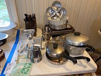 Overall view of the kitchen items, including pots, pans, Rival meat grinder, and serving tray on table.
