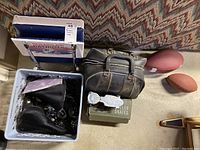 Photo showing vintage black Sears roller skates in a plastic bin, AMS bowling ball case with bowling shoes, Patriot blue folding stadium seat, and two brown footballs on the floor.