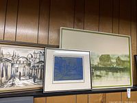 Three framed wall art pieces displayed on a wooden wall, including one large abstract landscape, one blue cityscape print, and one monochrome cityscape painting.