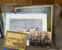 Six framed seascape paintings stacked against a wall, showing variety of maritime scenes including ships, sailboats and harbor activity.