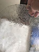 Close-up of clear glass platter showing embossed pine cone, twigs, and bow design details on the rim