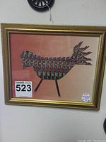 Full view of framed bird artwork made from cigar bands, showing the whole piece in gold frame on pink background.
