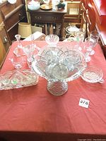 Clear pressed glass items arranged on a red cloth including pedestal punch bowl with cups, salt sellers, celery dish, nappy, cream and sugar set, candlesticks, mini compotes, and stemmed water glasses.