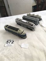 Four HO scale train items including clear locomotive, blue/gray engine, gray box car and TEXACO tanker car on white tablecloth with auction tag.