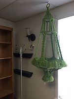 Full view of large green macrame plant hanger hanging by a metal ring from ceiling corner, showing wide open design, fringe details and size compared to adjacent coat rack and wall sconces.