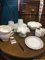 Photo showing various milk glass items on a wooden table including pedestal bowl, cups, creamers, and lace edge plate