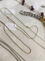 Photo showing gold-tone thinner chain necklaces laid on cream fabric background, also showing part of silver-tone beaded necklace and beaded necklaces with pendants.
