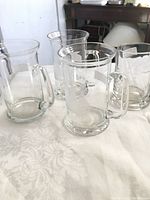 Four clear glass mugs arranged on a white patterned cloth. Three mugs are identical with large handles and cylindrical shape. Fourth mug shown on right has different handle and shape.