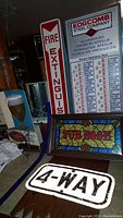 Photo showing the four vintage signs grouped on a table: Fire Extinguisher metal sign, Edgecomb Steel Company metal sign with specifications, wooden framed stained glass style Pub Room sign, and a white metal 4-Way sign.