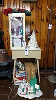 Photo shows vintage ceramic Christmas tree on a small table, next to boxed Santa figure and folded fabric tree skirts stacked on top. Includes string lights and additional ceramic figurines on the floor.