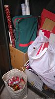 Photo showing multiple rolls of red, white, and patterned wrapping paper, green and red wrapping paper caddy, white plastic bag containing cellophane wrap, and cardboard boxes with gift boxes inside.