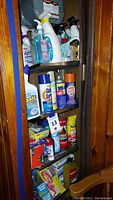 Wooden shelving unit with various cleaning spray bottles, disinfectants, wipes, and household liquids stacked on multiple shelves.