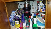 Photo showing under-sink area with various cleaning supplies including wipes, gloves, paper towels, and trash bags surrounding the garbage disposal.