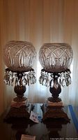 Pair of two crystal prism table lamps with spherical cut glass shades and decorative hanging prisms, sitting on brass/gold ornate bases with white marble squares.