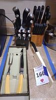 Photo showing multiple cutlery sets in blocks with black handles, meat carving set with yellow bakelite handles in box, and other utensils on counter.