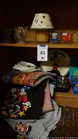 Vintage Disney-themed mugs including Tigger, Beauty and the Beast mug shaped like Mrs. Potts with a gold spout handle, and Garfield mugs alongside assorted Disney sweatshirts folded on a shelf.
