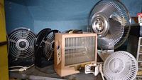 Photo showing multiple fans and an electric heater arranged on a shelf. The fans include a box fan, oscillating fans, and the heater is visible with controls.