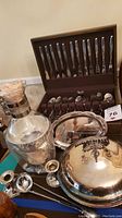 Photo showing stainless steel flatware in wooden box, silver plate serving items including lidded dish, candle holders, ice bucket with tongs.