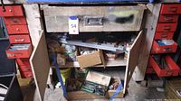 Photo of open metal workbench cabinet with shelves holding miscellaneous hardware, boxes, and tools.