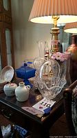 Photo showing various glass and ceramic decorative items arranged on wooden table near lamp, including cut glass vases, blue candy dish, small white ceramic bottle with floral bird motif, and a round crock.