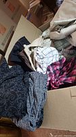 Cardboard box filled with assorted women's clothing including patterned pants and tops in various colors including black with swirl pattern, pink plaid, striped pants.