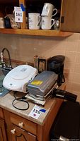 Photo of kitchen countertop with George Foreman grill, white pizzelle maker, black coffee maker, electric carver, and handheld mixer visible.