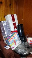 Photo showing boxed Gevalia coffee maker, boxed Melitta coffee maker, black 1-cup coffee maker, and boxed electric ice shaver on brown chair with wooden wall background.