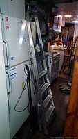 Full view of aluminum step ladder leaning beside a refrigerator and other basement items.