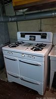 Vintage white Caloric gas range with 4 burners on top, two oven doors, two storage drawers below, and front control knobs in basement setting.