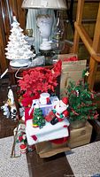 Vintage ceramic white Christmas tree with red bulbs and star topper, sitting on a wooden table beside lamps and poinsettia decorations. Includes Santa Santa figurine and small green Christmas tree in burlap base.