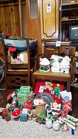 Wide view of vintage Christmas decorations including ceramic figurines on chair and numerous smaller items on floor including Christmas lights, candles, ornaments, poinsettia, stockings, and boxes.