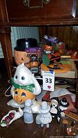 Wide angle photo showing a large ceramic pumpkin ghost figure, smaller pumpkin figures with cat themes, multiple ceramic pilgrim-style figures, and part of the wooden noisemaker on a wooden surface.