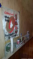Photo showing vintage Ortlieb's metal banner sign on wall and a row of 11 Bicentennial beer cans plus other assorted empty beer cans on shelf below.
