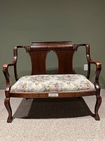 Front view of the solid wood antique bench showing floral upholstered seat and curved arms with carved floral detail on backrest.
