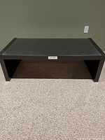 Front view of a black coffee table with glass top on a beige carpeted floor against a green wall.