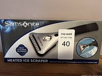 Box front showing Samsonite branding and image of the heated ice scraper. Features like melting ice and snow on contact, built-in flashlight, expandable handle, and power cord length are visible.