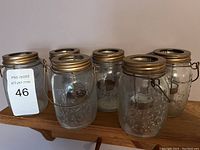 Front view of six empty glass Mason jar lanterns with gold-colored metal lids and handles arranged on a wooden shelf, showing embossed floral designs on the glass.