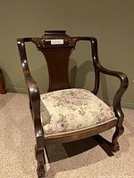 Full front view of the solid wood rocking chair with floral cushion on carpeted floor against a light olive wall.