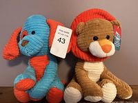Photo of two plush stuffed animals brand new with tags, one blue and orange dog and one brown lion with an orange mane.
