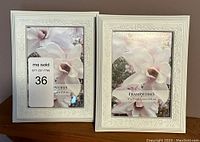 Two white plastic picture frames with floral embossed decorative edges, holding marketing inserts showing a white magnolia flower.