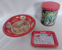 Three Coca-Cola themed tins including a wide shallow bowl, small rectangular tin plate, and tall round canister with red lid.