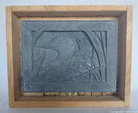 Gray carved relief sculpture of two birds among reeds displayed in a wooden box, photo taken from an angle showing side and top.